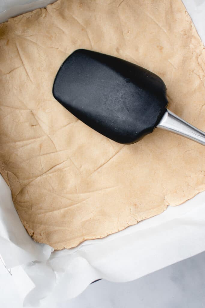 A black silicone spatula spoon smooths the crust dough into an even layer, in a parchment lined baking pan.