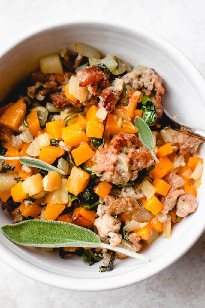A white bowl with butternut squash hash, garnished with fresh sage leaves.