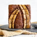 loaf of sliced banana bread stacked vertically on a round slate serving platter on top of a burlap sack. there is a stainless steel fork to the right of the loaf of bread