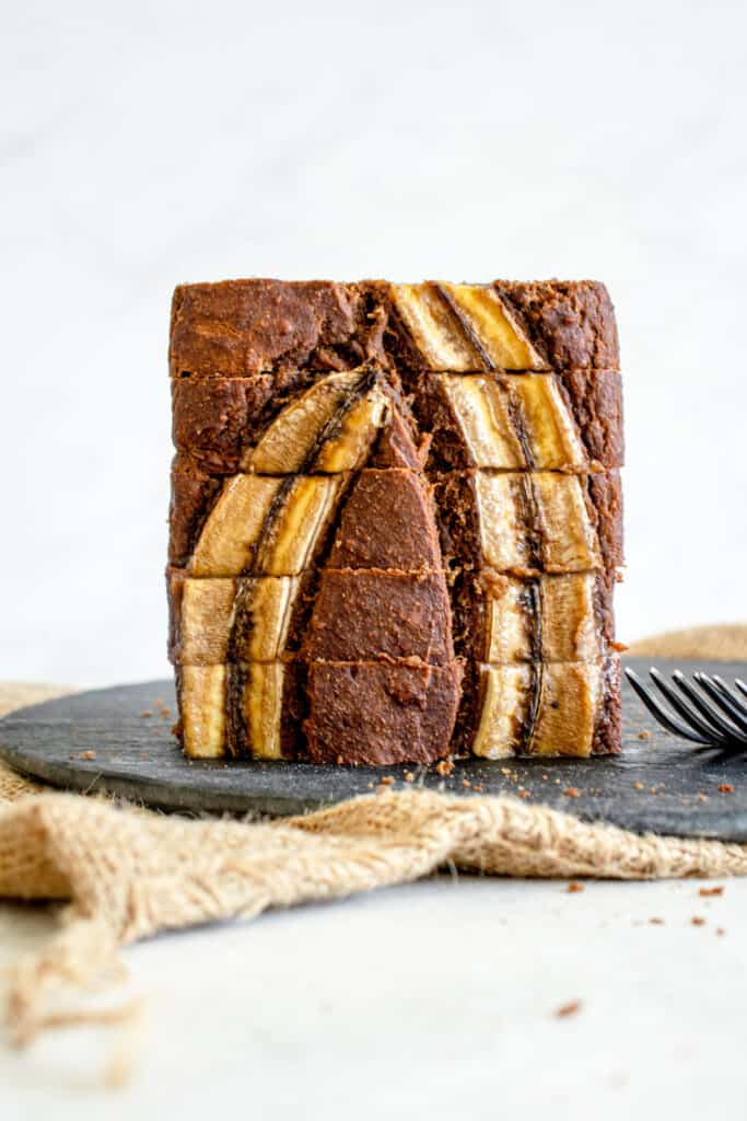 loaf of sliced banana bread stacked vertically on a round slate serving platter on top of a burlap sack. there is a stainless steel fork to the right of the loaf of bread