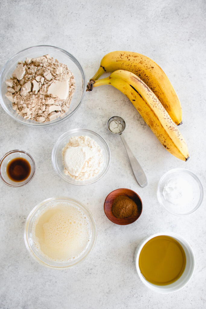 overhead shot of ingredients for the paleo banana bread including white bowl with olive oil, clear glass bowl with gelatin egg, small wooden bowl with cinnamon, small glass bowl with vanilla extract, small glass bowl with baking soda, measuring spoon with salt, glass bowls with coconut flour and green banana flour and two ripe bananas with brown spots