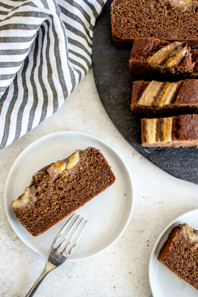 two small white plates with a slice of banana bread on each plate. there is a fork resting on one of the plates and a large round slate grey platter with additional slices of banana bread in the top right of the image