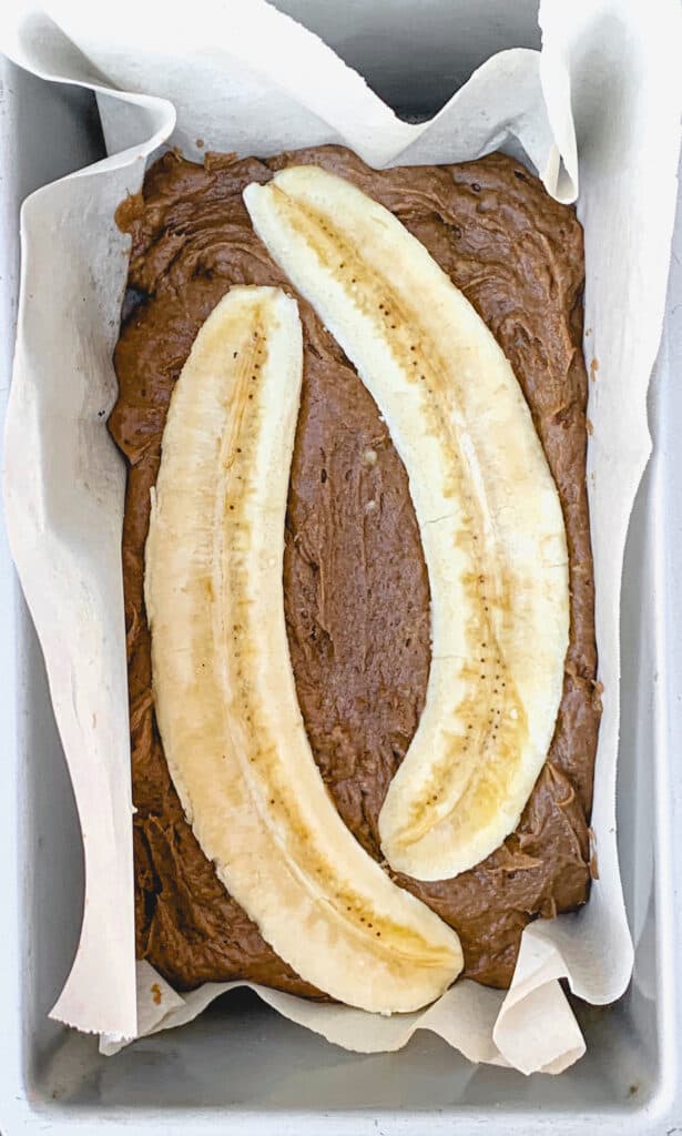 overhead shot of banana bread batter in parchment lined baking sheet with one banana sliced vertically and opened in half on top the batter
