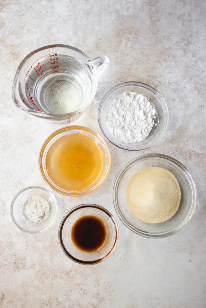 Overhead shot of small glass bowls with measured out ingredients to make Gluten-Free Marshmallows. Pictured clockwise from top: arrowroot starch, gelatin, vanilla extract, salt, honey, and a glass measuring cup with water.