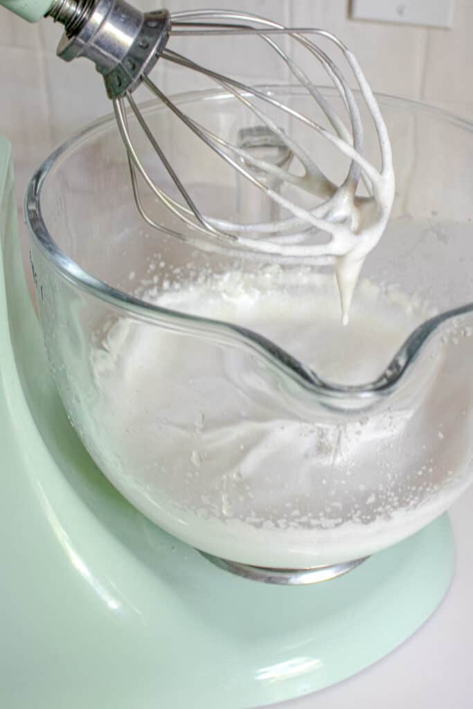 A stand mixer with gluten-free marshmallow mixture in the glass bowl, with the whisk attachment raised out of the bowl with the mixture dripping from it.