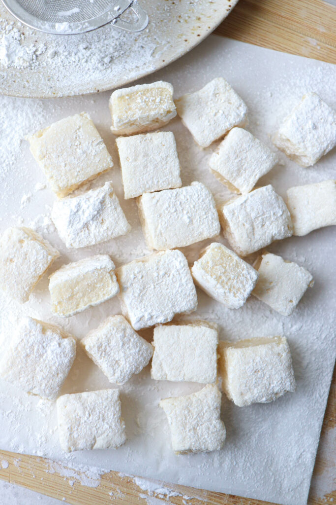 Overhead shot of a piece of parchment paper with large Gluten Free Marshmallows dusted with arrowroot starch, on top of a wooden cutting board. In the top left corner, there is an off white plate speckled with brown spots with a a partial sieve shown, and some arrowroot starch sprinklings.