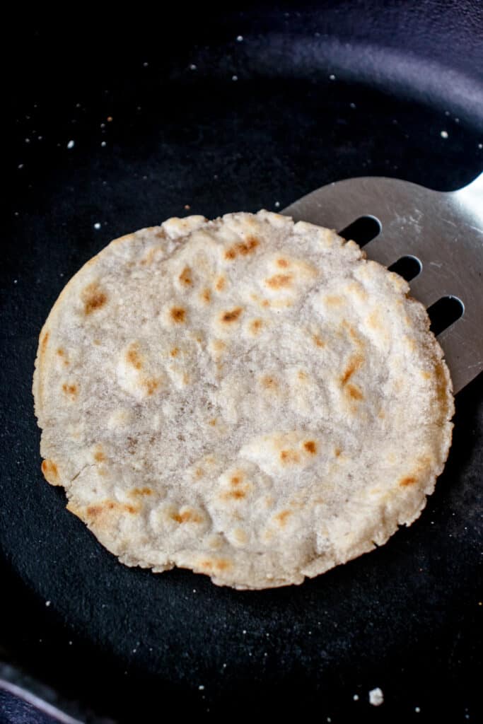 A cooked Gluten Free Cassava Flour Tortilla with some brown spots on the surface in a cast iron pan, with a spatula lifting it out.