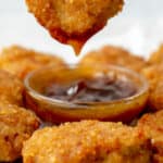 A brown hand dipping a golden brown chicken nugget into a small glass bowl of brown sweet and sour sauce surrounded by chicken nuggets, on parchment paper.