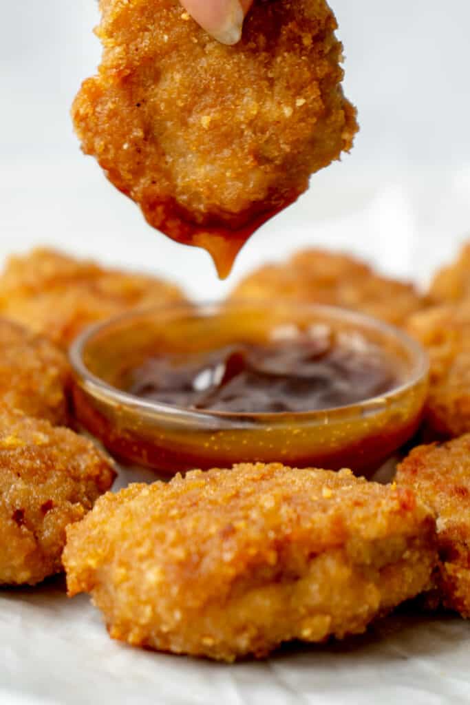 A brown hand dipping a golden brown chicken nugget into a small glass bowl of brown sweet and sour sauce surrounded by chicken nuggets, on parchment paper.