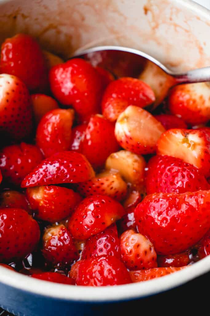 Strawberries stewing in the lemon sugar mixture in a saucepan, with a spoon.