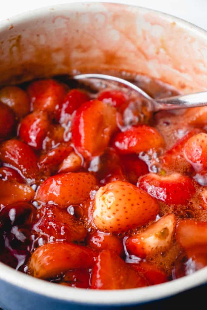 Strawberries simmering in the strawberry lemon maple sugar mixture to make strawberry compote, in a saucepan with a spoon.
