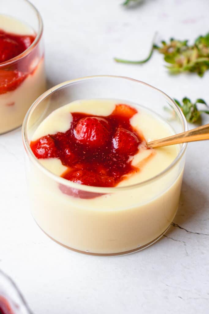 Close up of a short glass jar with lemon custard and strawberry compote with a gold spoon dipped in.