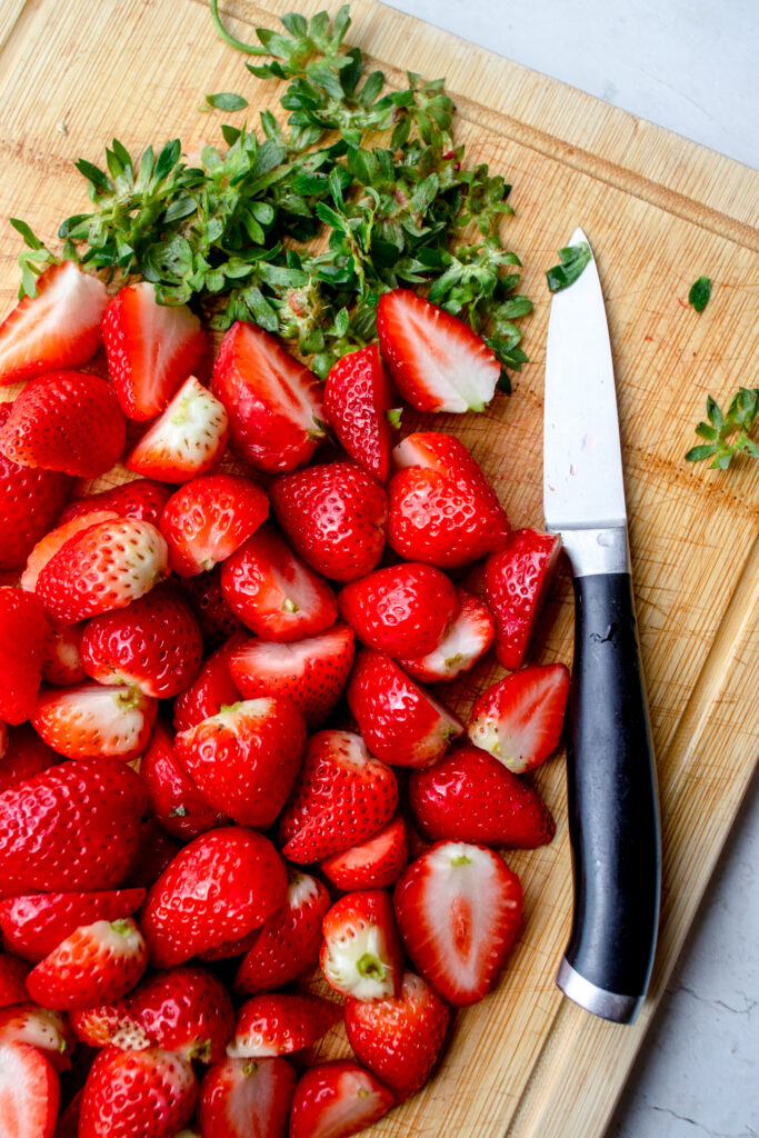 Strawberries cut in half with green stems cut off, on a wooden cutting board with a knife.
