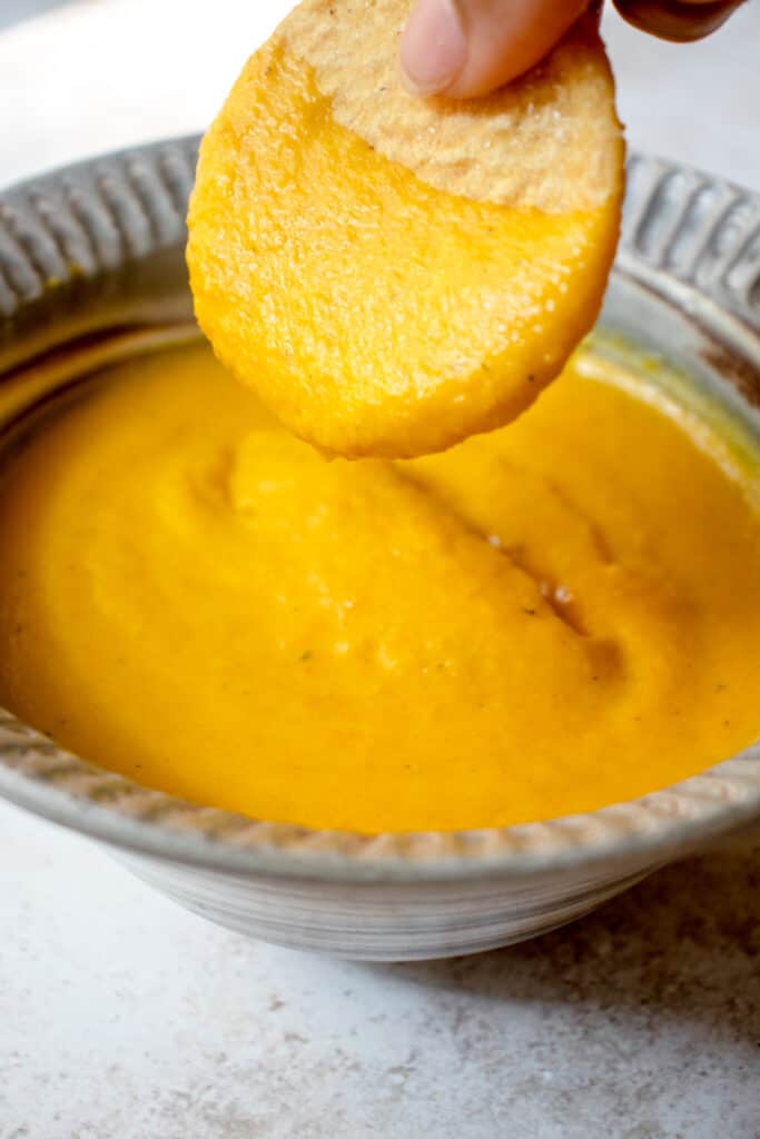 Brown hand holding a chip which has been dipped into the Vegan Queso Cheese Sauce below in a small serving dish.