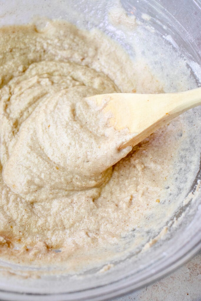 Dry and wet ingredients mixed together with a wooden spoon in a large glass mixing bowl to form a batter.