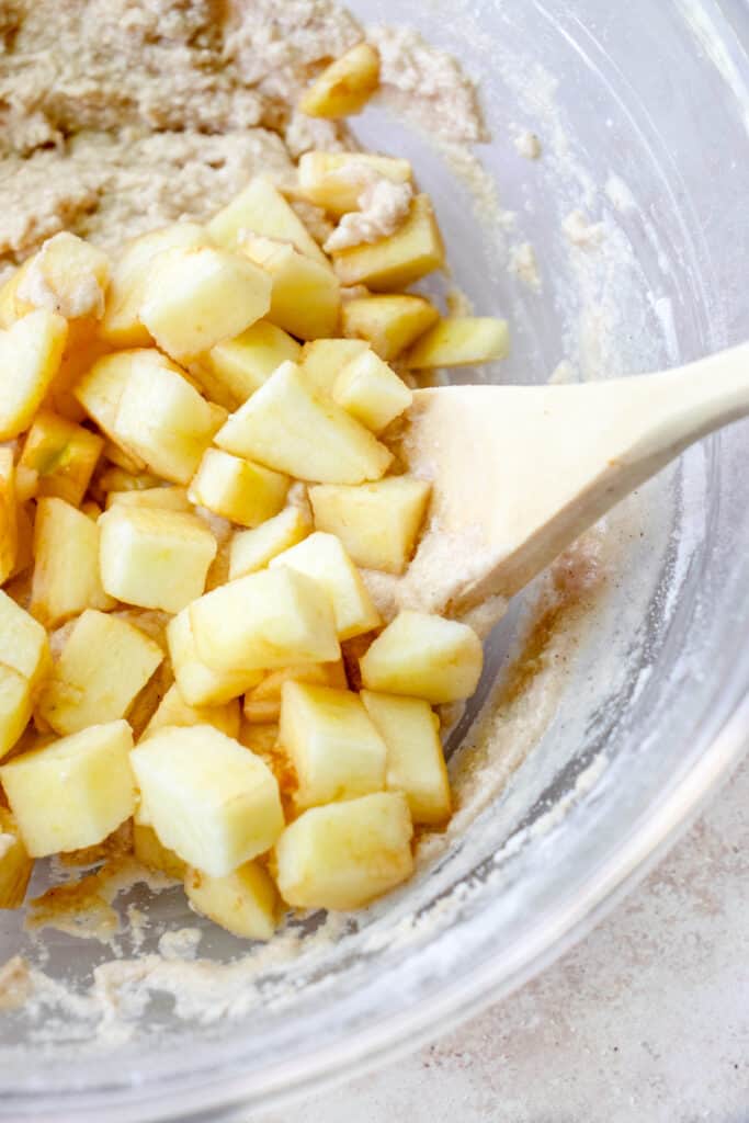 Peeled and diced apples added to the apple fritter batter with a wooden spoon in a large glass bowl.