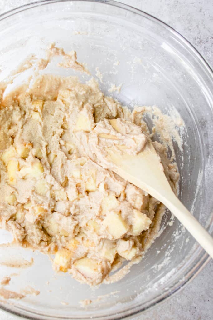 Diced apples folded into the apple fritter batter with a wooden mixing spoon in a large glass bowl.