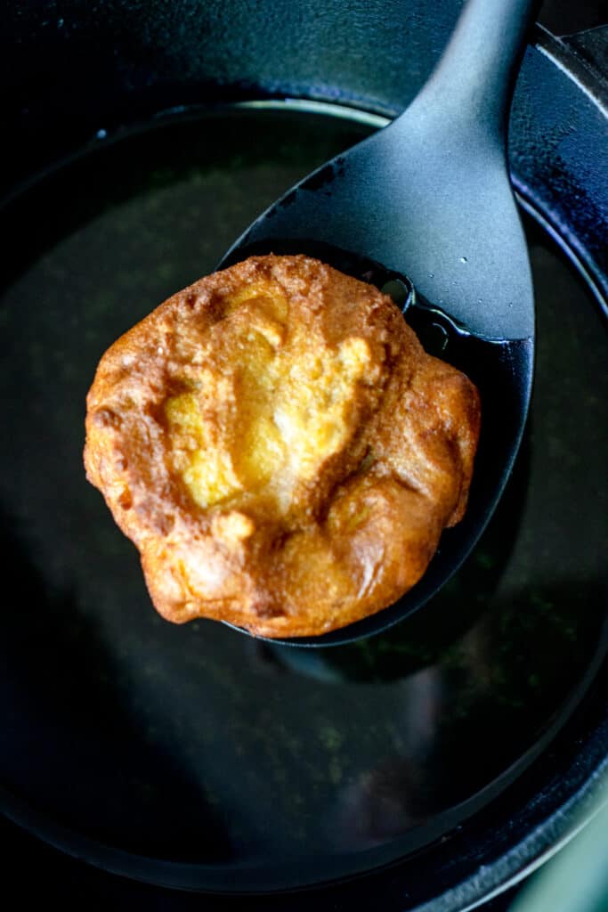 A slotted spoon scooping an apple fritter out of frying oil in a cast iron pot.