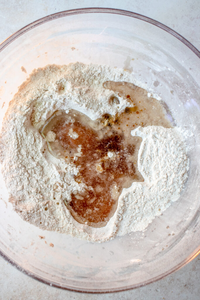 large glass bowl containing unmixed dry and wet ingredients for pumpkin sugar cookies. the wet ingredients of maple syrup, coconut oil, vanilla are sitting in the middle of the dry ingredients