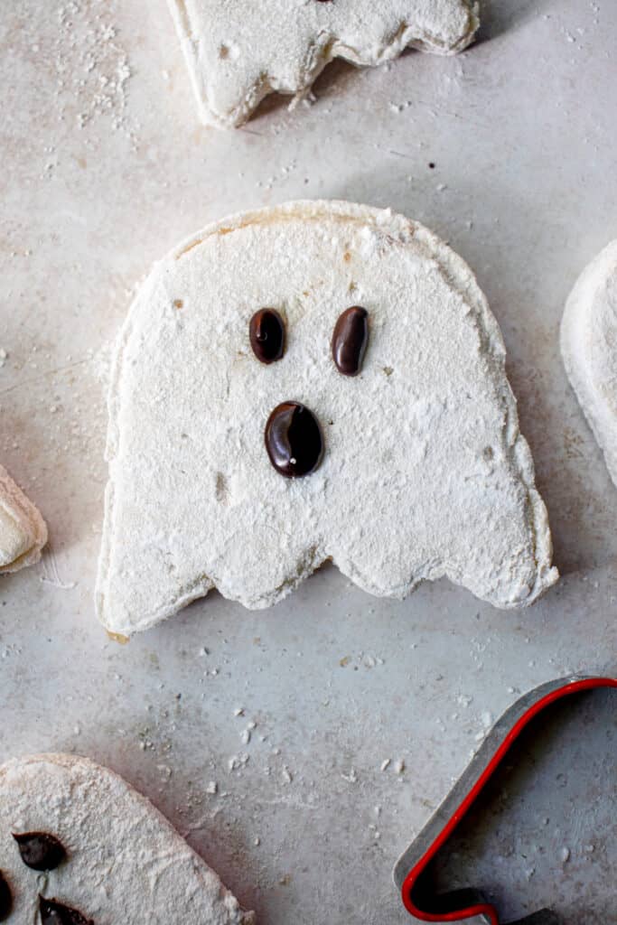 Ghost shaped vanilla marshmallow with brown eyes and mouth made of melted chocolate. To the bottom right of the frame is the red and stainless steel ghost cut-out mould.