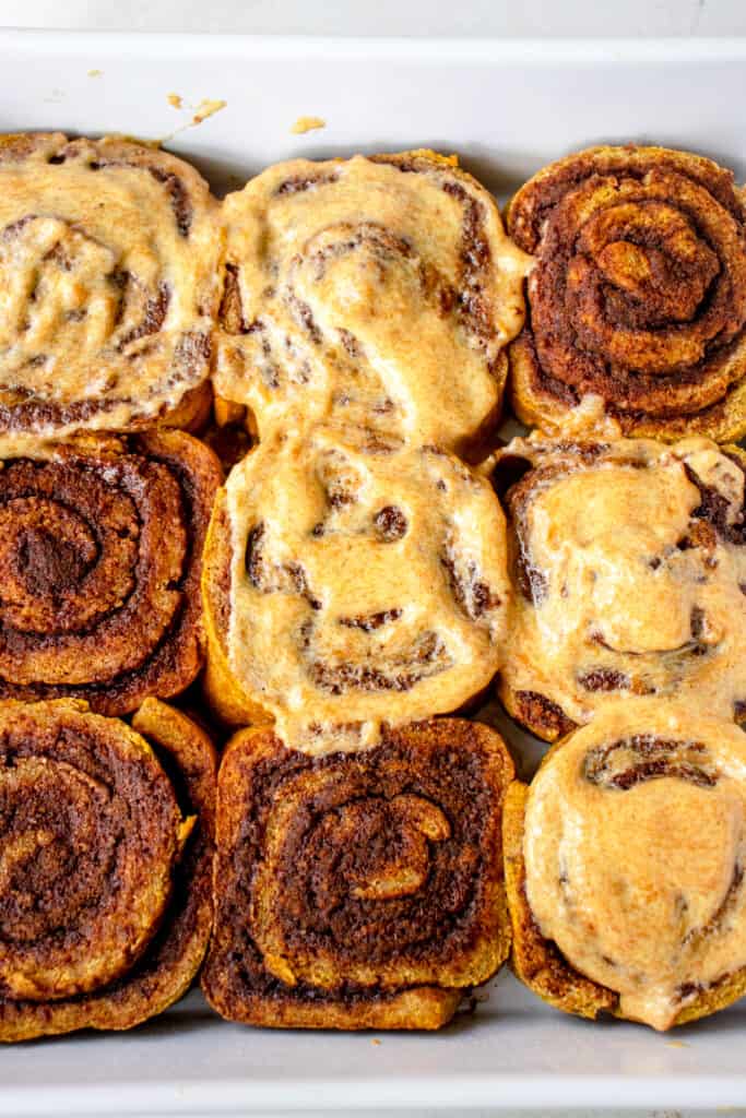 Baked Gluten-Free Pumpkin Cinnamon Rolls in a baking dish. Five of the nine rolls are iced with the Vegan Pumpkin 