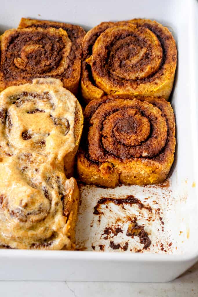 Close up of 5 baked Gluten-Free Pumpkin Cinnamon Rolls in a baking dish. Two are iced with the Vegan Pumpkin Buttercream Frosting.
