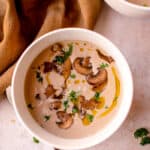 Gluten-Free Dairy-Free Cream of Mushroom Soup in two bowls. One bowl is in the center and the other is in the top right of the photo. The soups are garnished with a drizzle of olive oil, chopped fresh parsley and some ground black pepper. A brown linen napkin is gathered on the left side of the bowls, with a silver spoon placed on the left of the centered bowl.