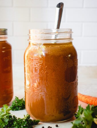 Two large glass mason jars with Gluten-Free Bone Broth. The jar in the foreground has a metal spoon in it while the jar in the background has a lid on it. The jars are surrounded by fresh herbs, a carrot, and whole black peppercorns.