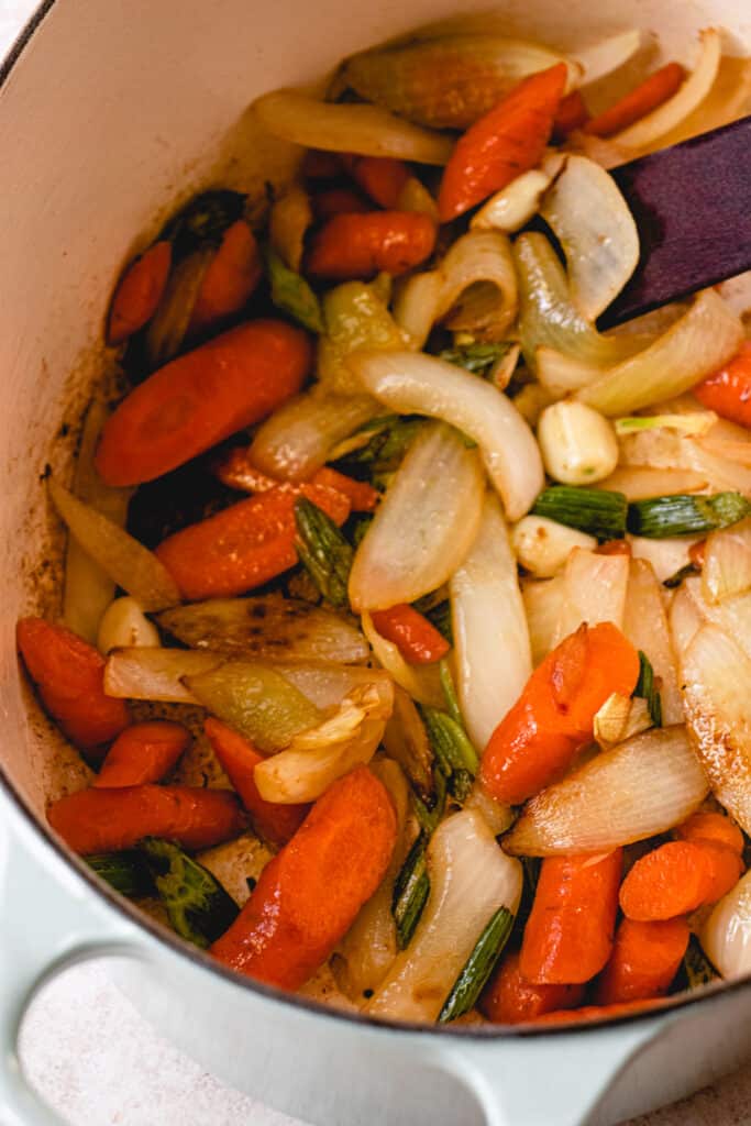 Caramelized, sautéed carrots, onion, garlic, and celery in a large pot with a wooden sauté stick.