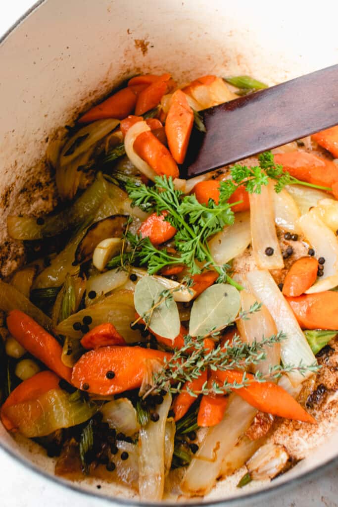 Large pot with sautéed vegetables, fresh herbs, and black peppercorns being stirred with a wooden sauté stick.