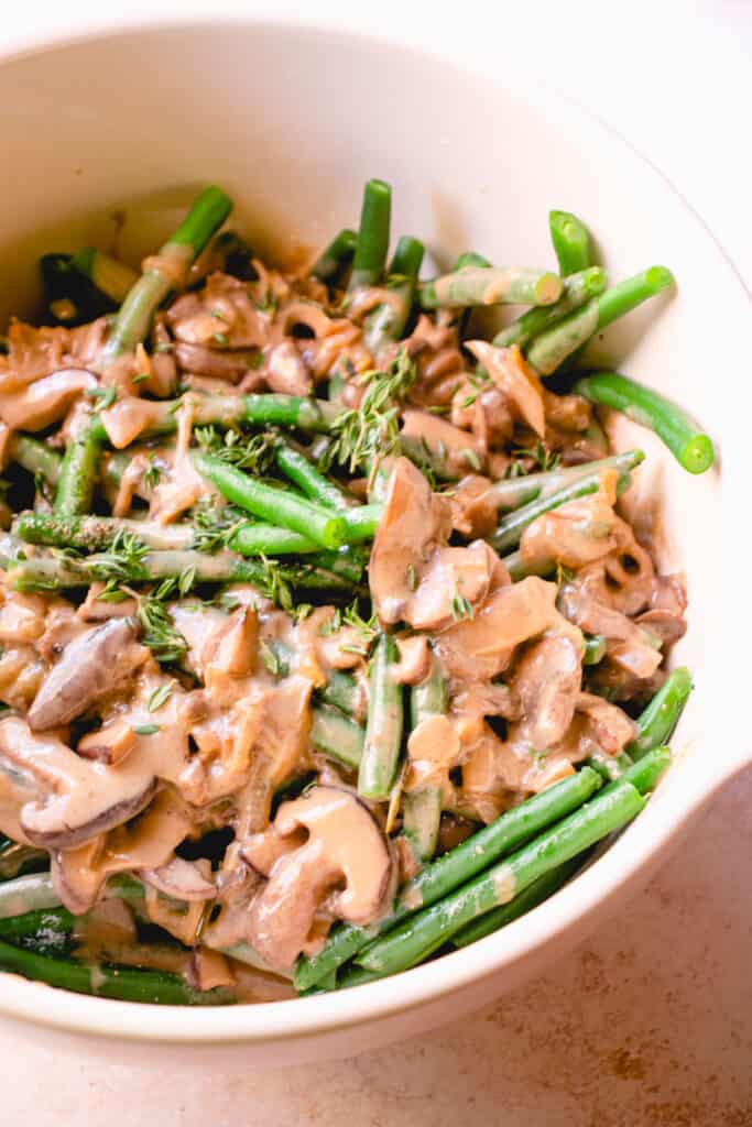 A large mixing bowl with blanched green beans, sea salt, black pepper, cream of mushroom soup and thyme leaves.