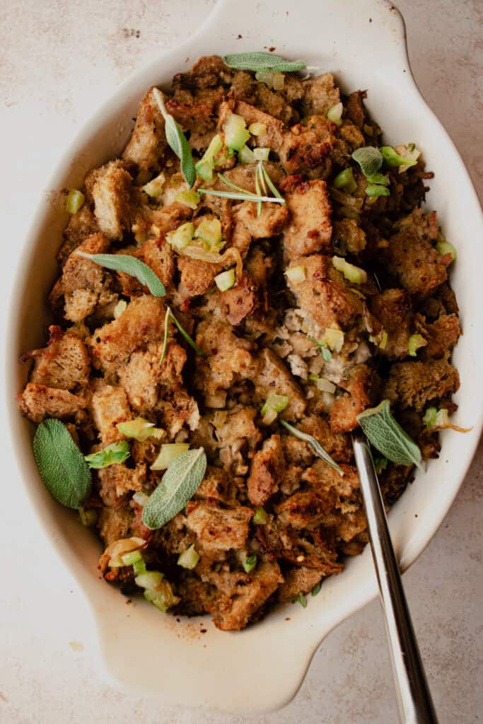 Gluten-Free Stuffing in a casserole dish garnished with diced celery and fresh herbs. A silver serving spoon is in the dish.