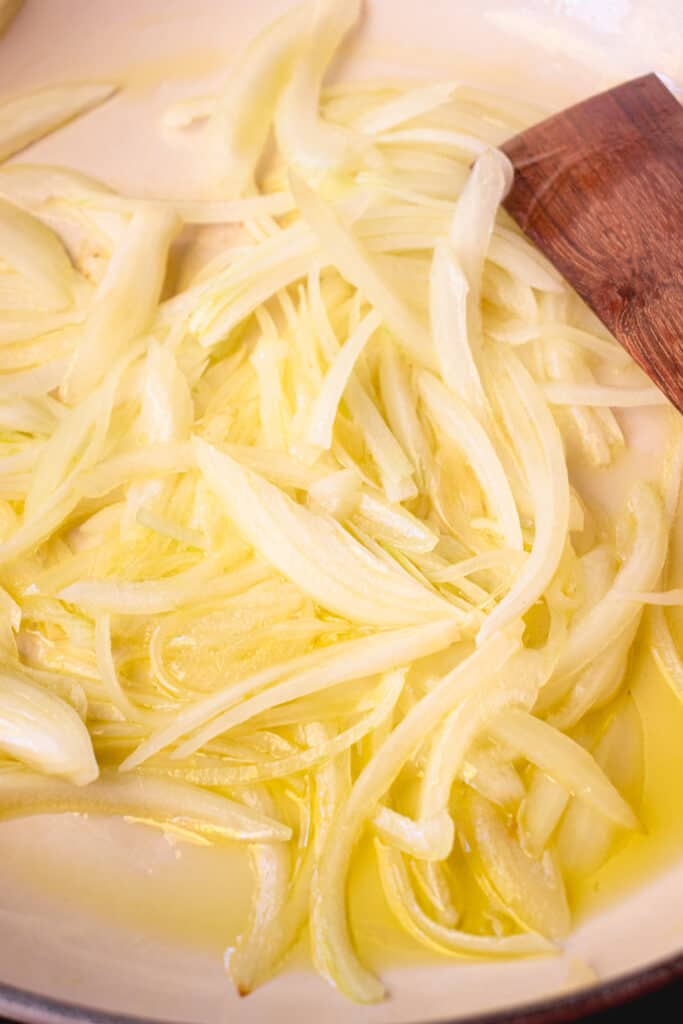 A skillet with olive oil, sliced onions, and a wooden stirring stick.