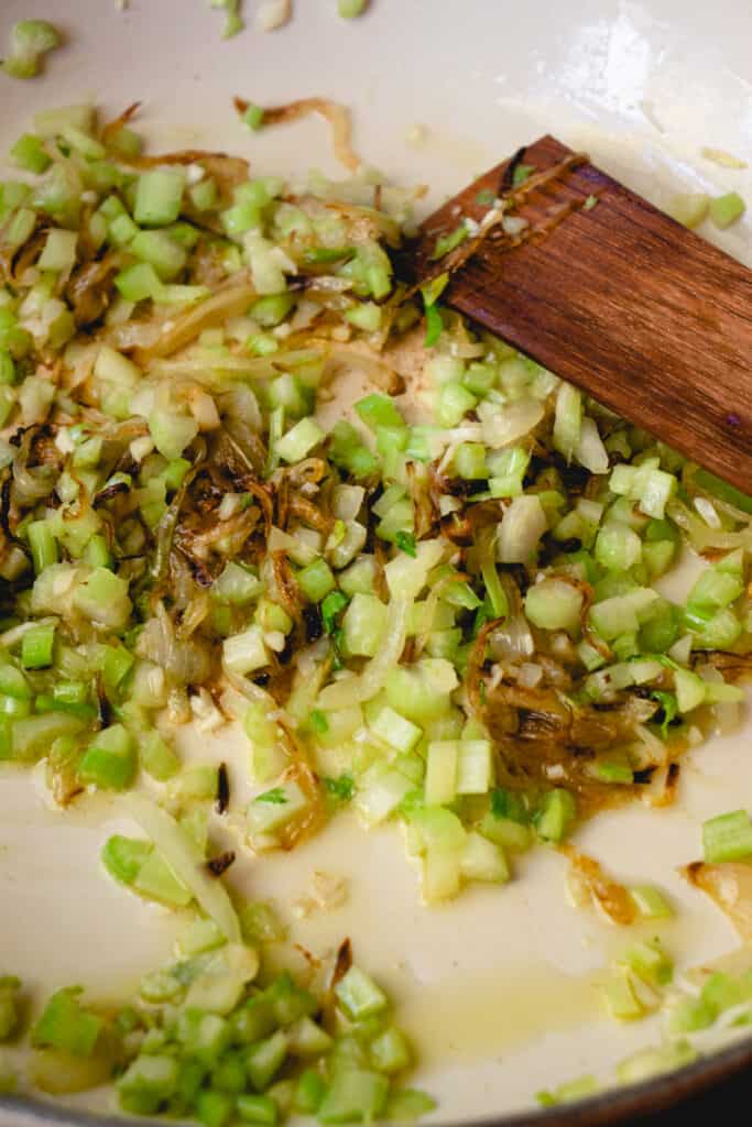 Caramelized onions with sautéed celery and garlic in a skillet being stirred with a wooden stirring stick.