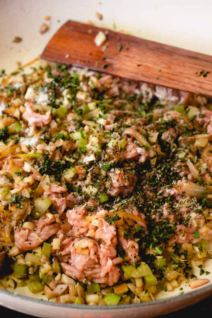 Ground pork and fresh herbs added to sautéed vegetables in a skillet. A wooden stirring stick rests in the top of the pan.