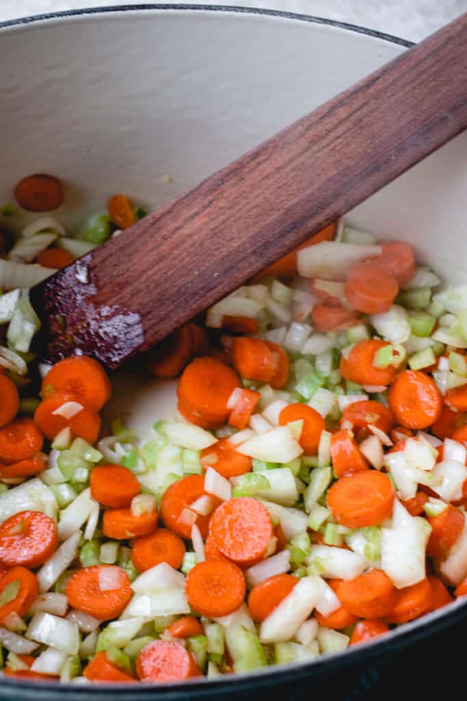 Carrots, celery and onions sautéeing in a pot with a flat wooden sauté spatula.