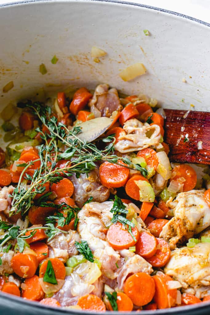 Herbs, spices, chicken thighs and vegetables cooking in a French oven and a flat wooden sauté stick in the pot.