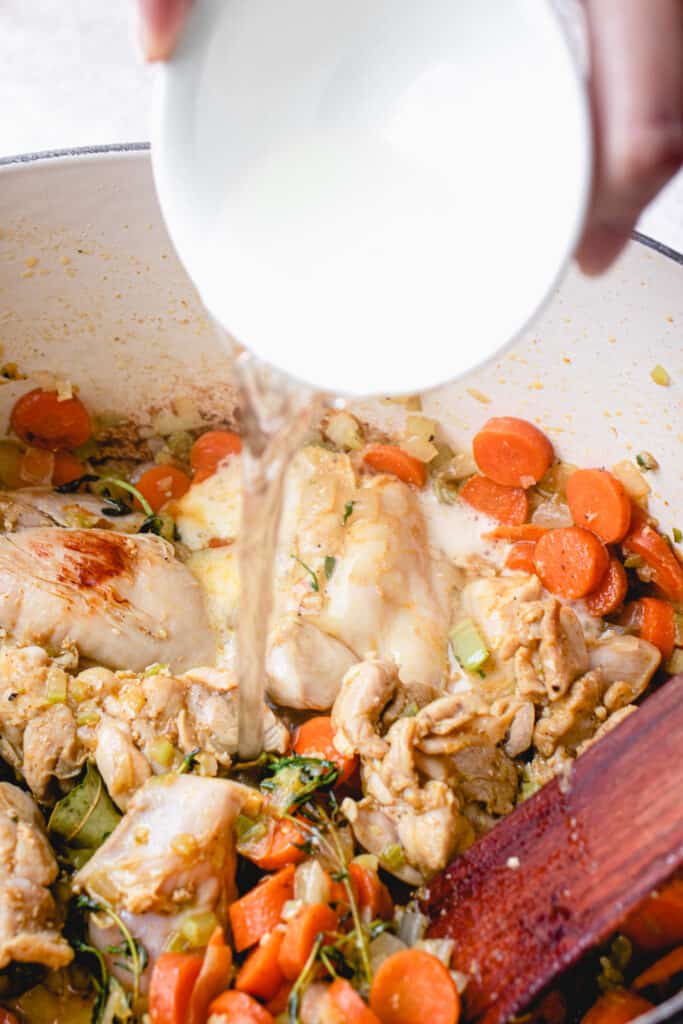 A brown hand pours white wine into the pot of chicken, herbs, spices, and vegetables.