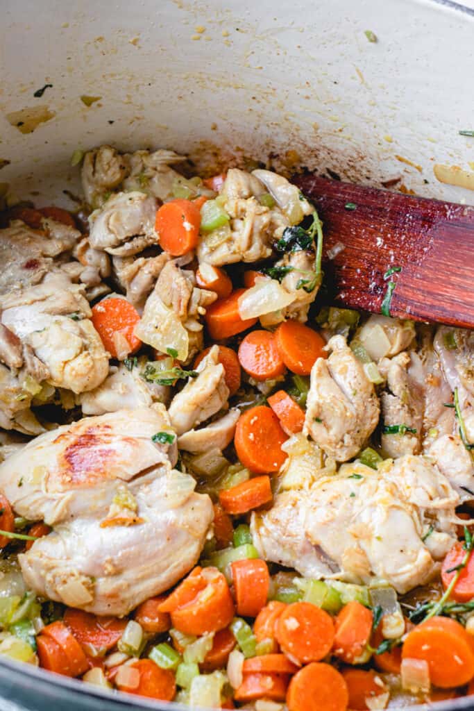 French oven with chicken, vegetables, herbs and spices, and a flat wooden sauté stick.