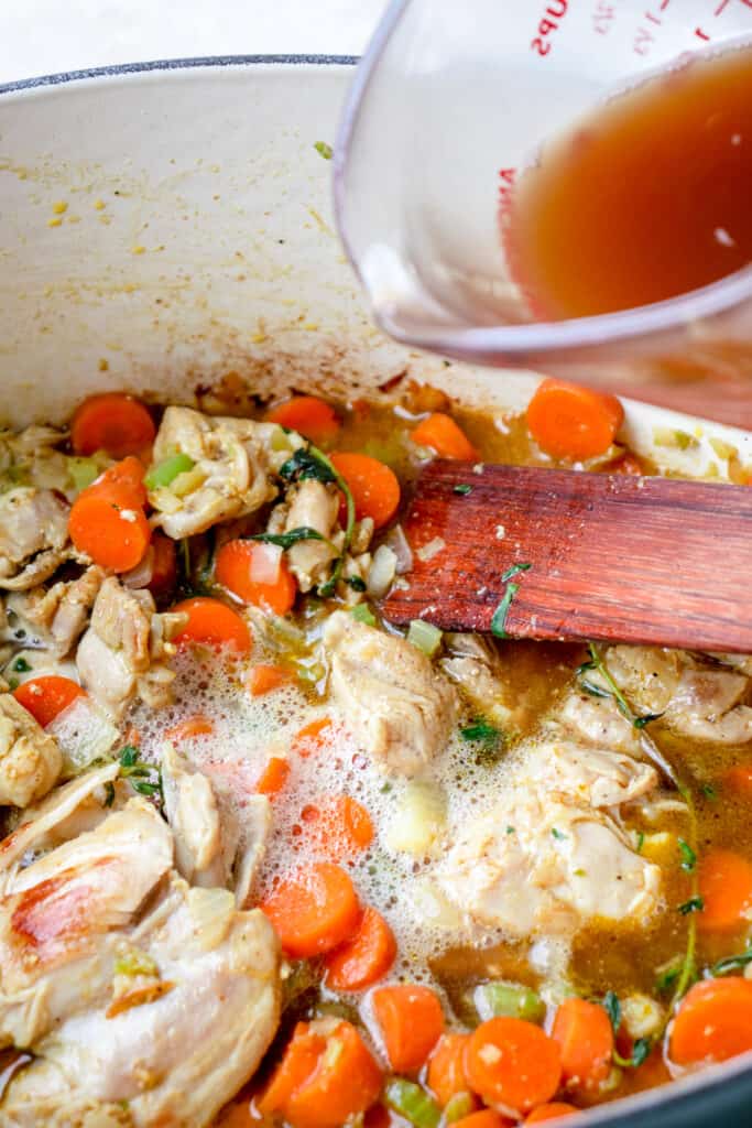 Chicken broth being poured into the French oven with chicken, vegetables, herbs and spices, and a flat wooden sauté spatula.