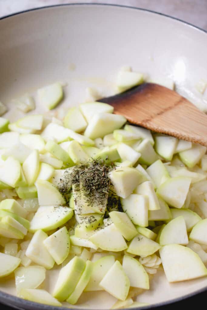 peeled and chopped zucchini sauteed in a pan with onions and garlic and thyme and a wooden spatula