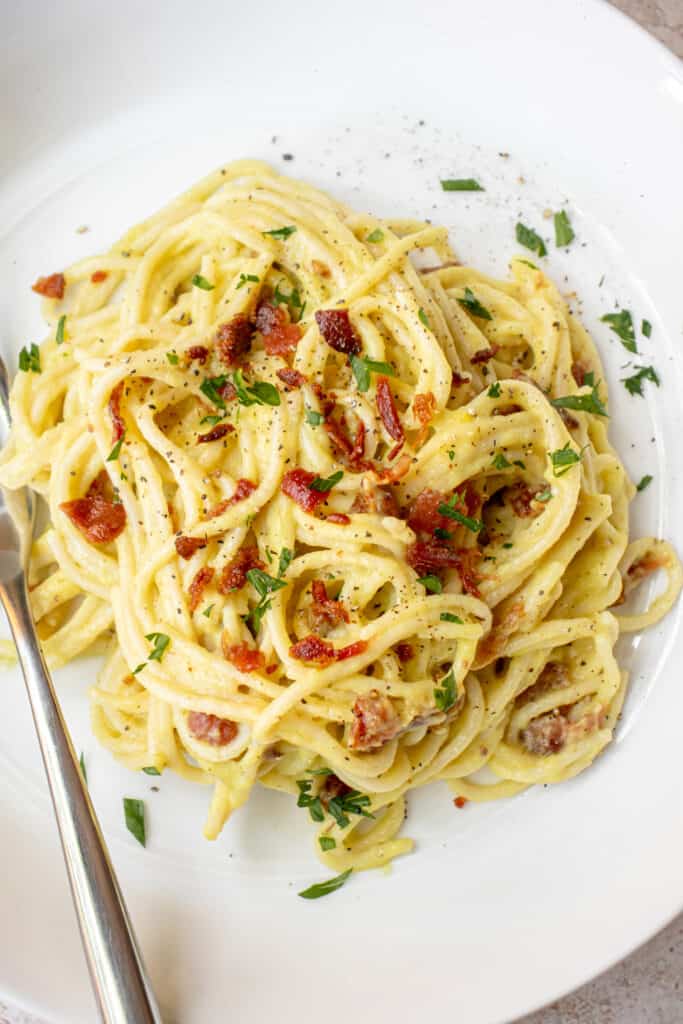 white plate with a stainless steel fork and pasta carbonara made of spaghetti with crumbs of bacon and garnished with parsley.