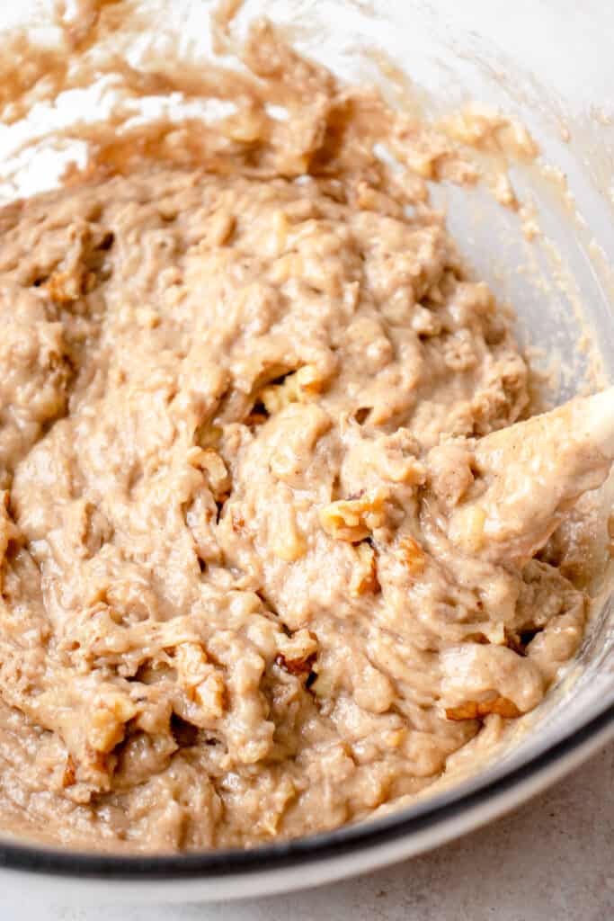 Banana bread batter in a glass bowl with a wooden spoon folding in walnuts.