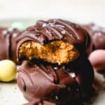 A stack of two DIY Vegan Chocolate Covered Easter Eggs with some more in the back and foreground, blurred, with colored mini eggs. The top cookie is bitten to show the interior texture of the filling.