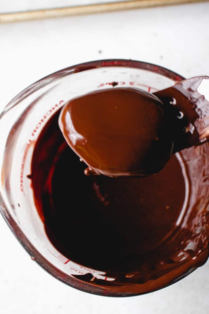 A fork holds an egg dipped in melted chocolate above a measuring glass with melted vegan chocolate.