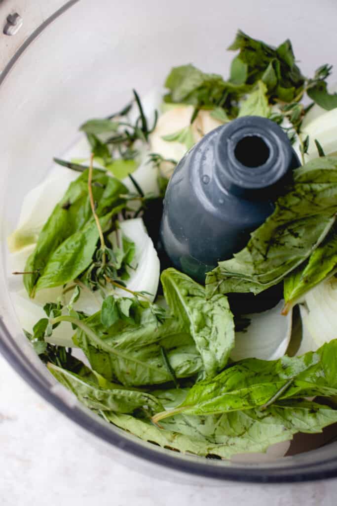 A food processor with fresh herbs, onion, garlic, and celery.