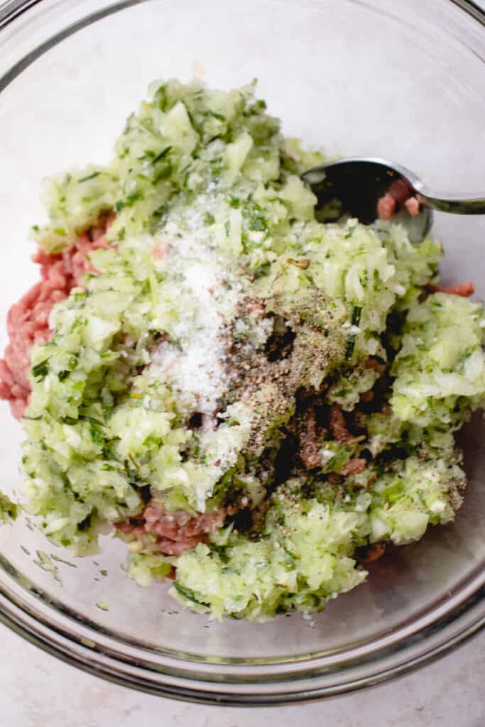 A glass bowl with ground beef, pulsed and processed herb and vegetable mixture, salt, and black pepper.