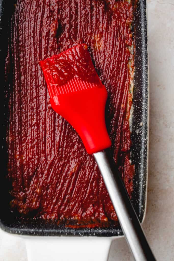 A rubber spatula brushes on the glaze atop the meatloaf mixture in a loaf pan.