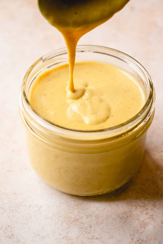 A spoon drips the Dairy-Free Cheese Sauce into a jar of it below.