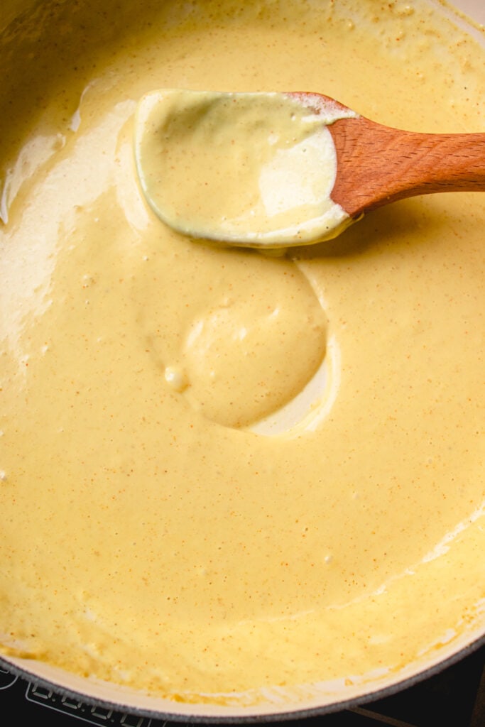 A wooden spoon swirls Dairy-Free Cheese Sauce in a skillet.